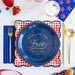 Napkin with 'Home of the Free, Land of the Brave' text on a blue plate with red checkered border, surrounded by gold cutlery and strawberries.