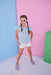Young girl in a light blue top and white shorts standing against a colorful background
