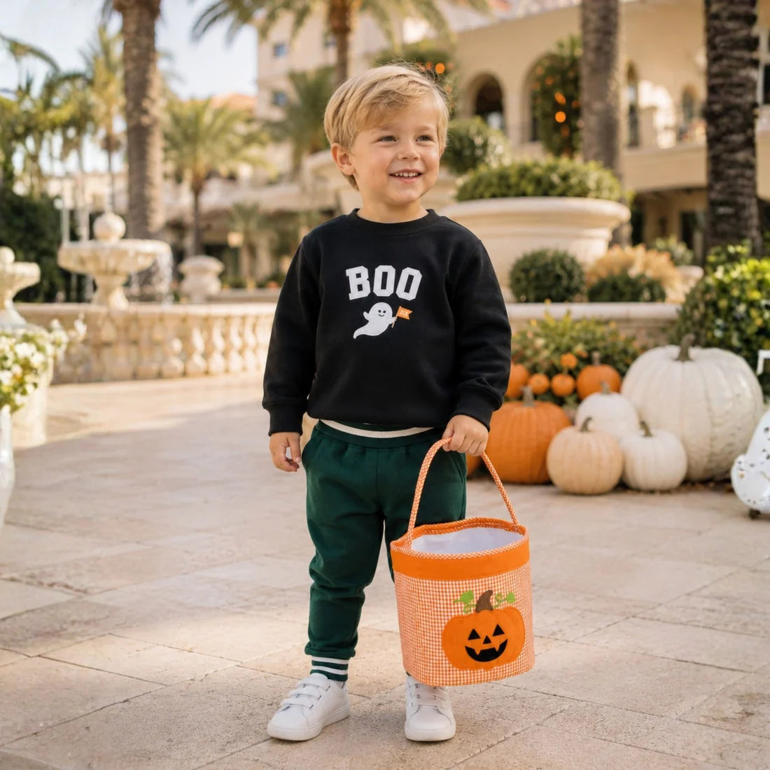 A young boy stands outdoors in a sunny courtyard holding an orange pumpkin-themed treat bucket. He is wearing a black sweatshirt with 'BOO' and a ghost graphic, dark green pants, and white sneakers.