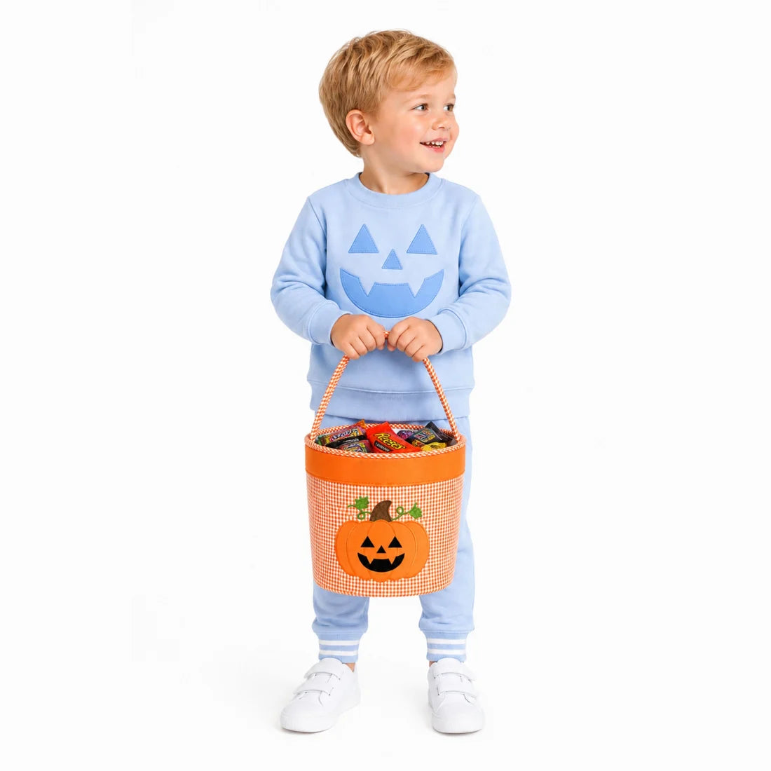 A young child wearing a light blue sweatshirt and matching pants stands against a white background holding an orange gingham Halloween candy basket with a jack-o'-lantern face.