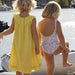Two children in yellow dress and floral swimsuit by a body of water.