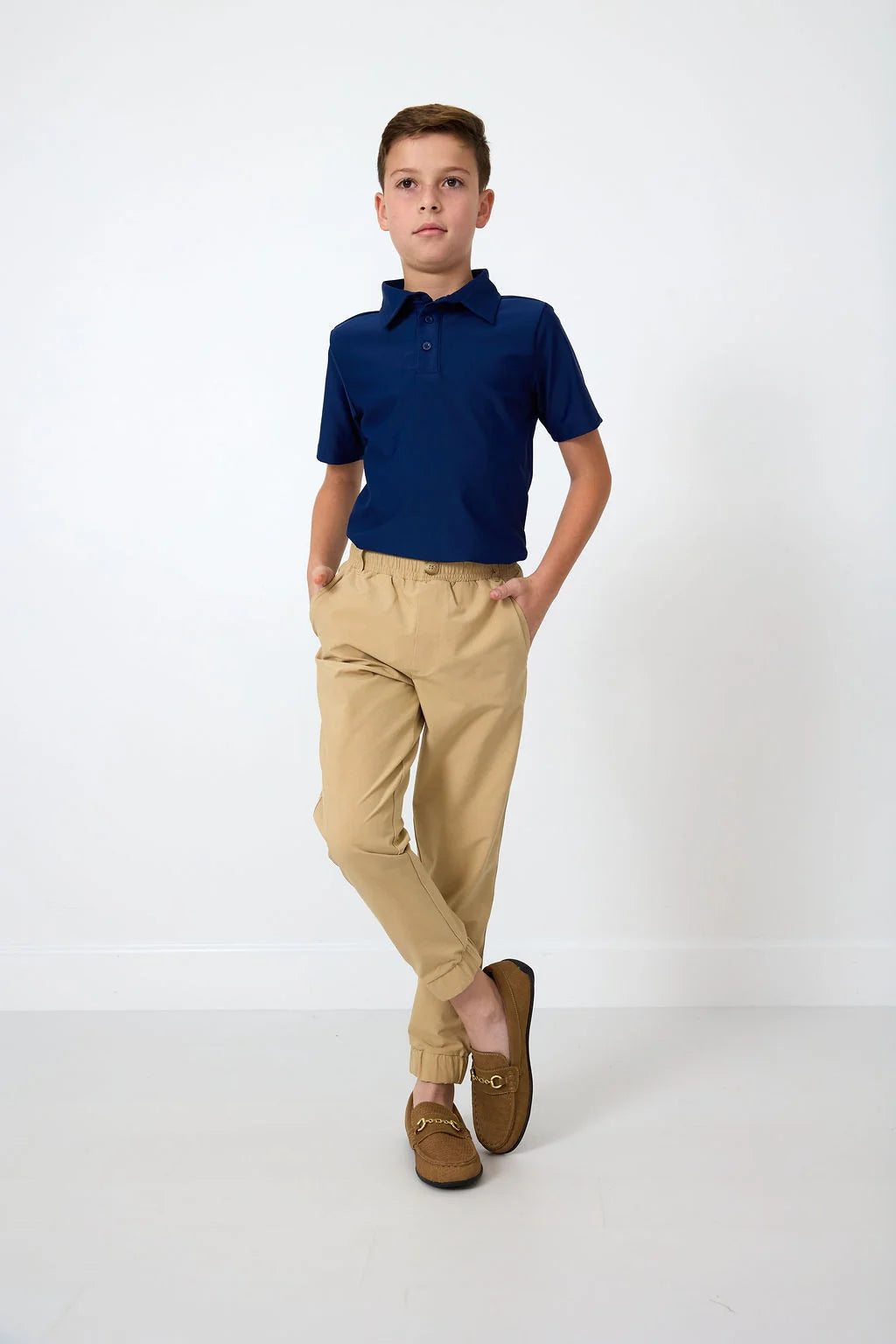 A young boy modeling a navy blue short-sleeve polo shirt and khaki joggers with brown loafers, standing against a clean white studio background.
