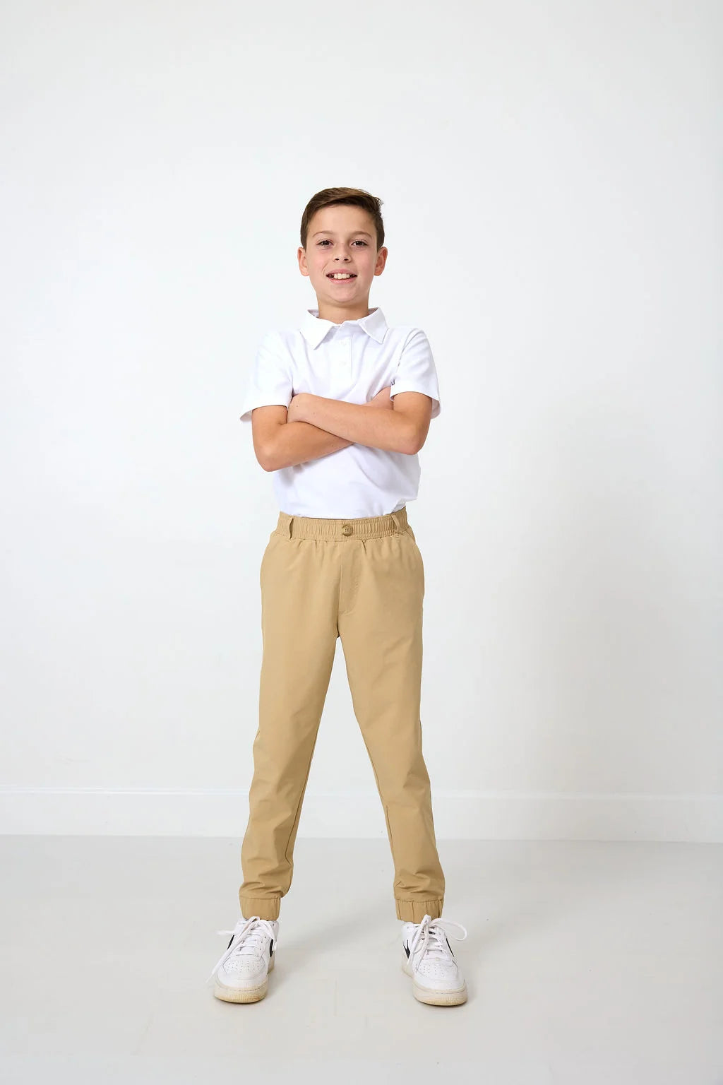 A young boy stands with arms crossed, wearing a white short-sleeve polo shirt, khaki joggers, and white sneakers, photographed against a clean white studio background.