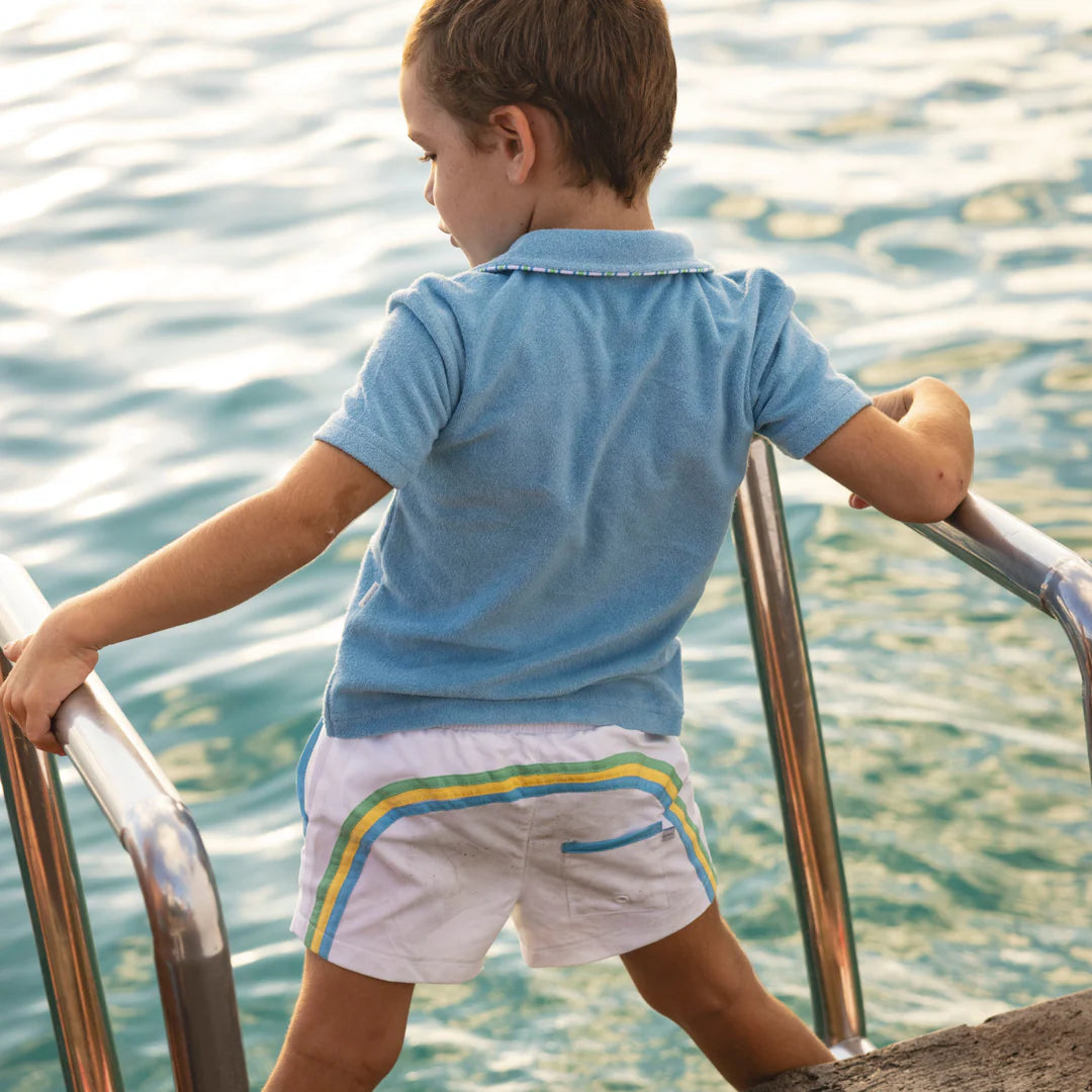 Child in a blue shirt and white shorts with colorful stripes standing by a body of water.