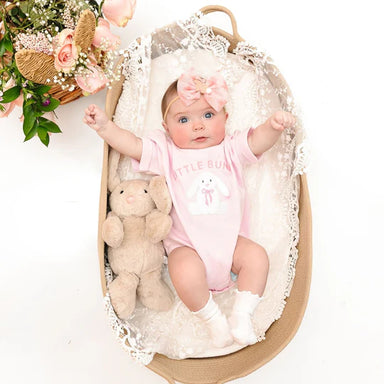 Baby in a pink onesie with bunny design, sitting in a decorative crib with flowers and a teddy bear.