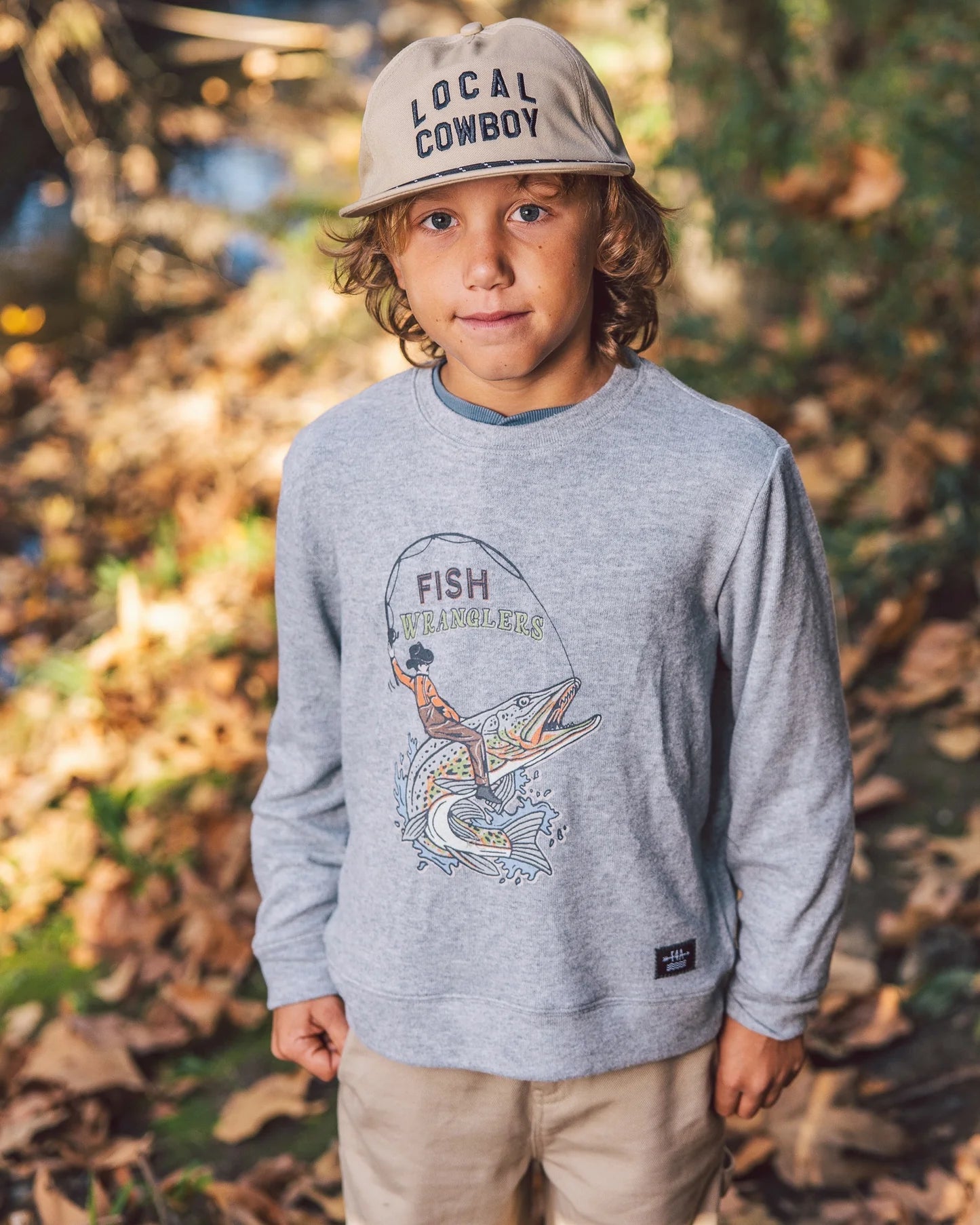A young boy wearing a khaki colored snapback hat featuring brim rope and embroidered "Local Cowboy" graphic on the front panel.
