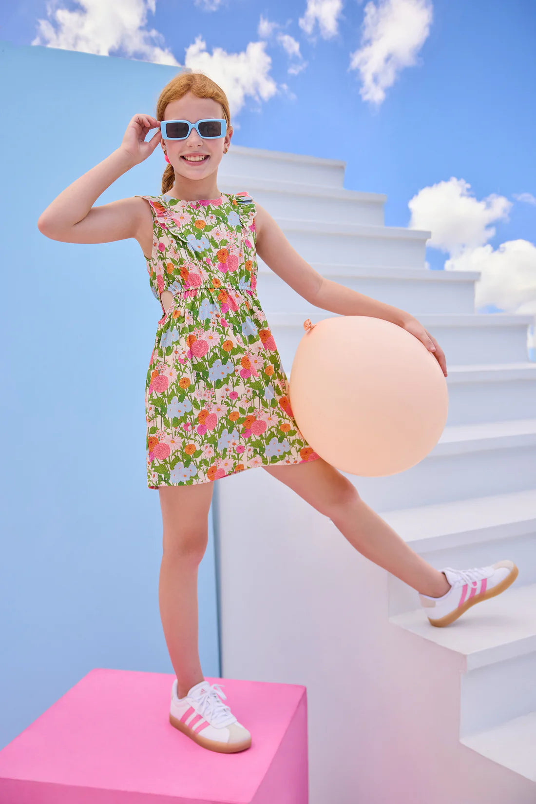Young girl in a floral dress standing on a pink platform with a white staircase and blue sky in the background.