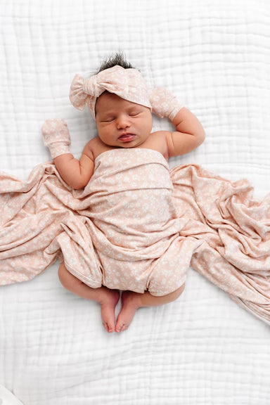 A sleeping newborn baby wrapped in a soft blush pink patterned swaddle blanket, wearing a matching pink knotted headband and mittens, lying on a white textured muslin blanket.