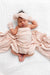 A sleeping newborn baby wrapped in a soft blush pink patterned swaddle blanket, wearing a matching pink knotted headband and mittens, lying on a white textured muslin blanket.