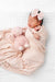 A sleeping newborn baby wrapped in a soft blush pink patterned swaddle blanket, wearing a matching pink knotted headband and mittens, lying on a white textured muslin blanket.