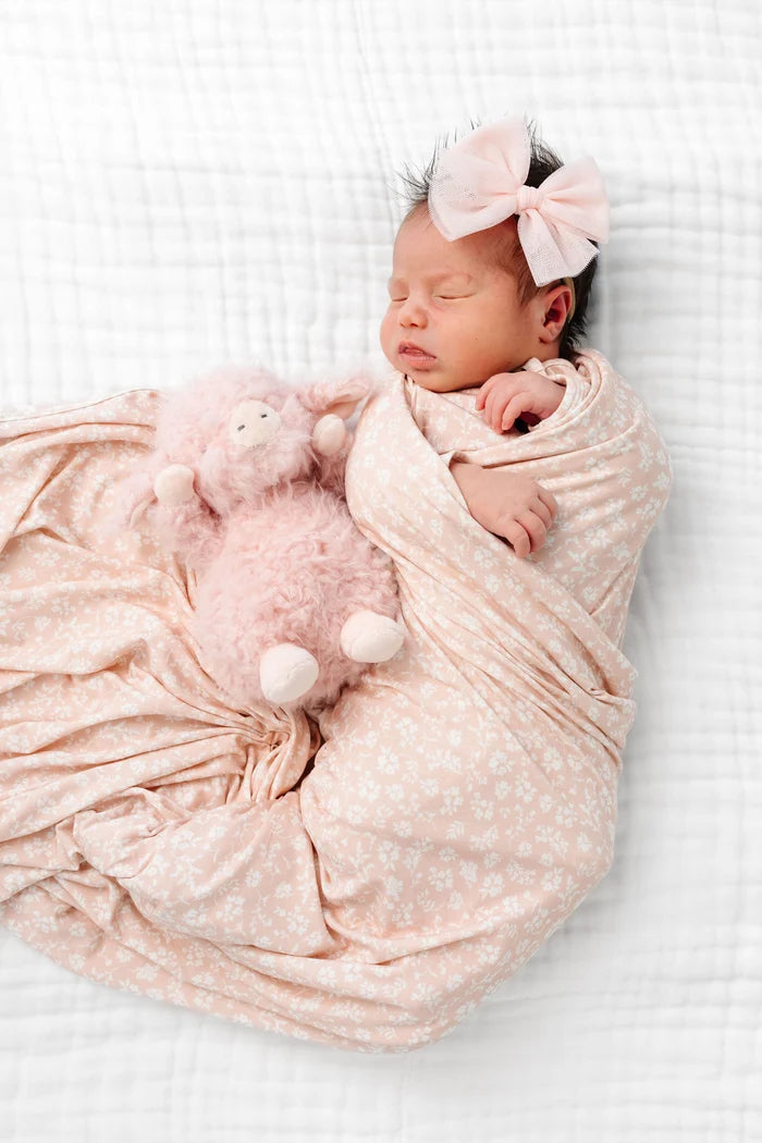 A sleeping newborn baby wrapped in a soft blush pink patterned swaddle blanket, wearing a matching pink knotted headband and mittens, lying on a white textured muslin blanket.