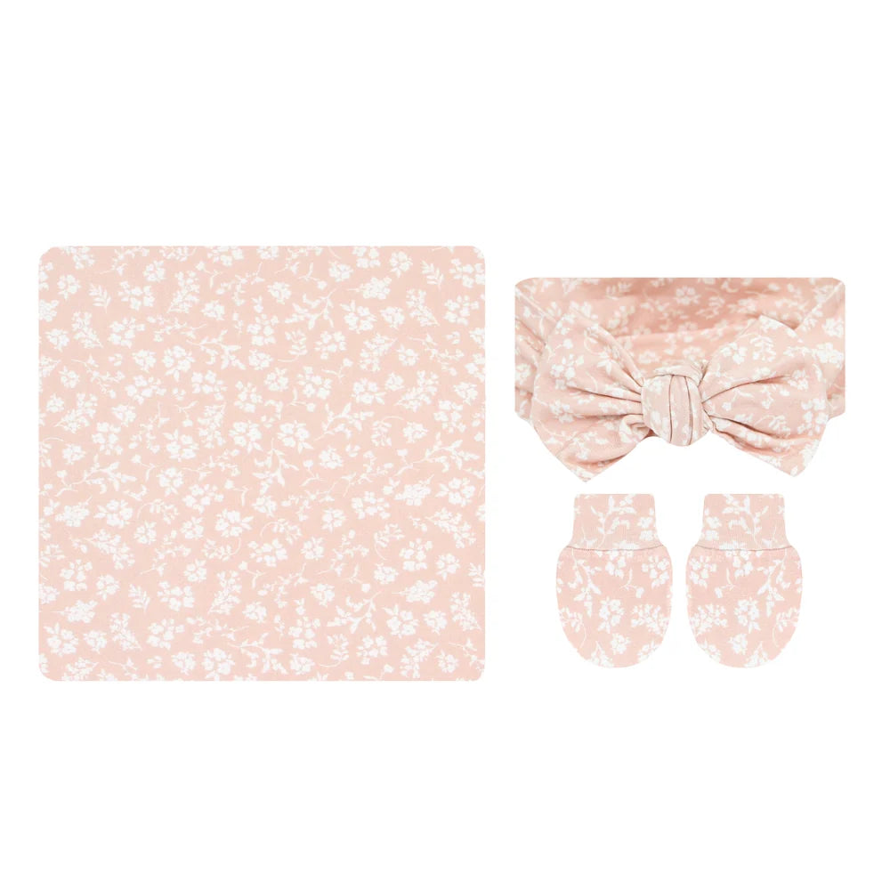 A baby accessory set featuring a square blanket, a matching bow headband, and a pair of mittens, all in a soft peach-pink color with a delicate white floral pattern, displayed on a solid white background.