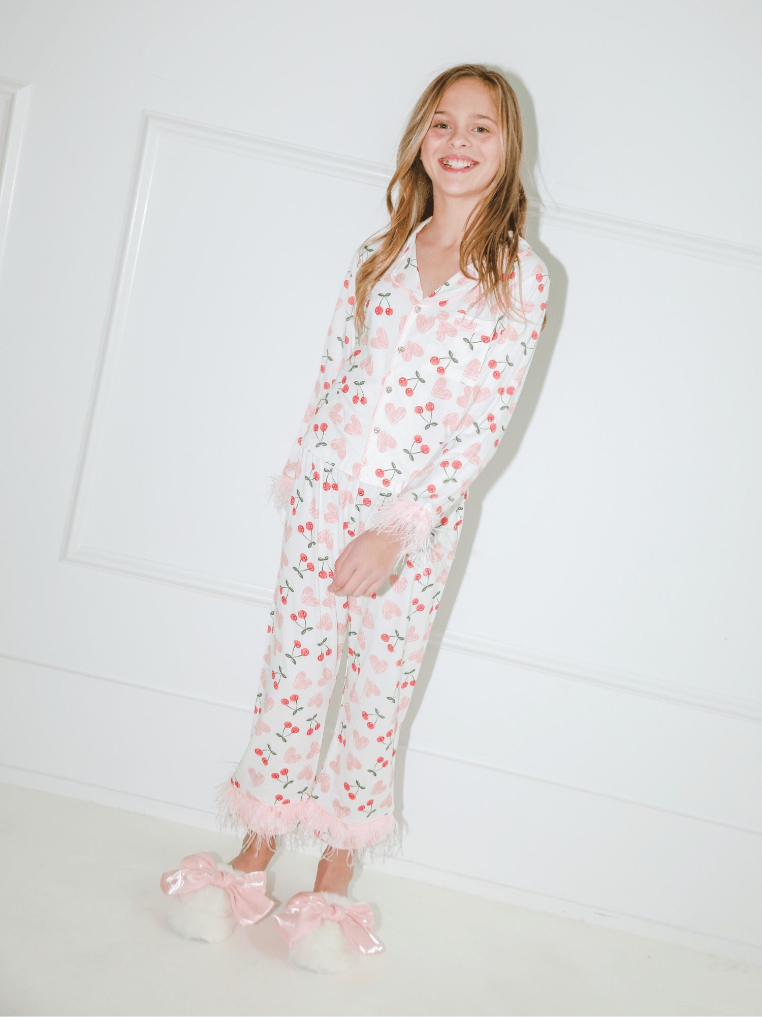 A young girl smiling while wearing a long-sleeve white pajama set with a pink cherry print and fringed trim, paired with fluffy white slippers with pink bows, standing against a white paneled wall.