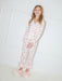 A young girl smiling while wearing a long-sleeve white pajama set with a pink cherry print and fringed trim, paired with fluffy white slippers with pink bows, standing against a white paneled wall.