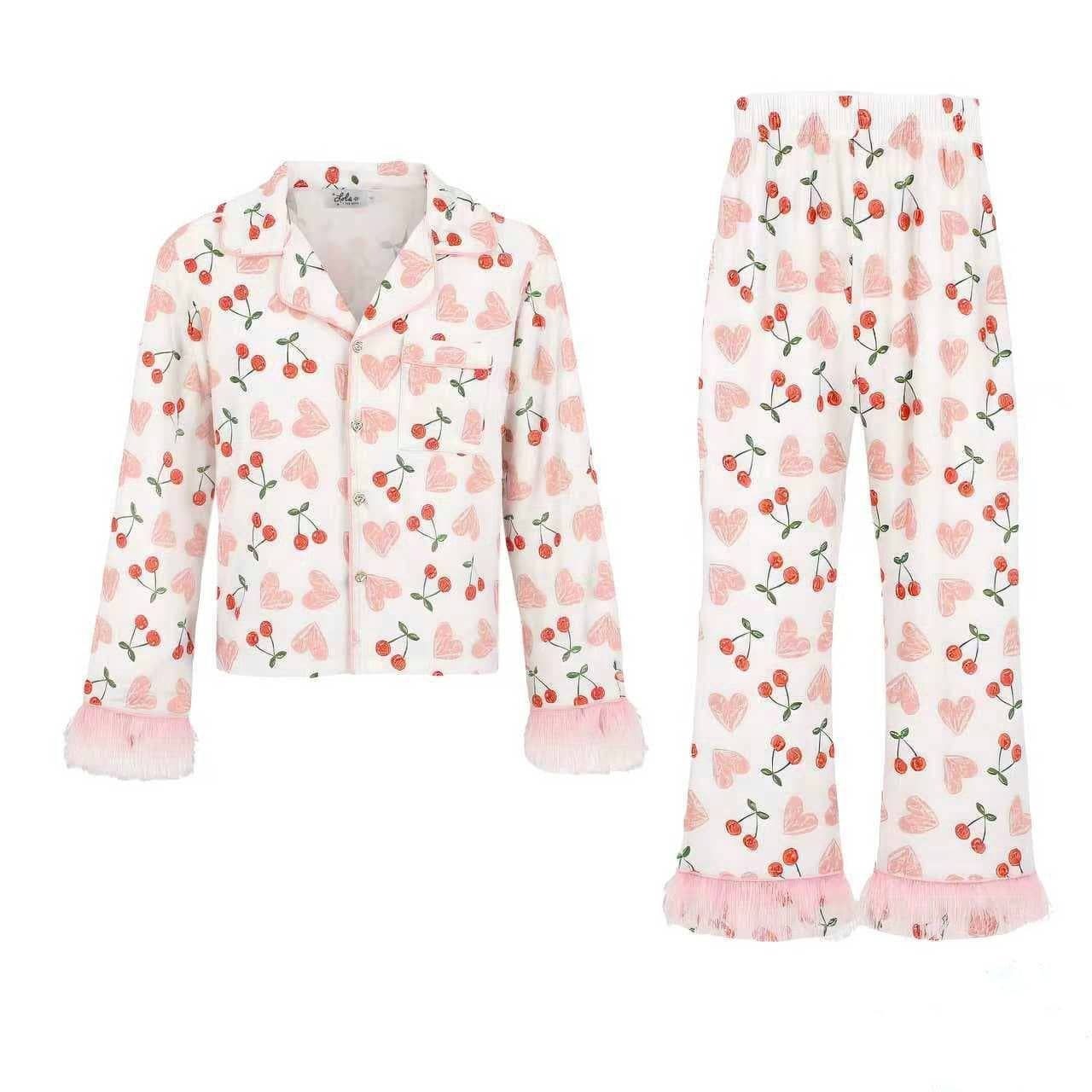 A two-piece girls' pajama set featuring a long-sleeve button-front top and matching wide-leg pants, both in white with a pink cherry and heart pattern and pink ruffled trim.