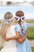 Two children wearing headbands with bows, one in a white dress and the other in a light blue dress, standing close together outdoors.