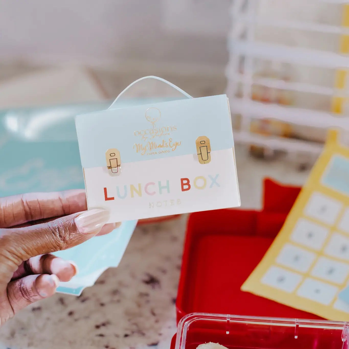Hand holding a small lunch box with a decorative label, blurred background