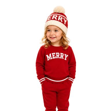 A young child wearing a festive red and cream knit holiday set, including a sweatshirt, pants, a knit pom-pom beanie, and a matching belt, standing against a solid white background.