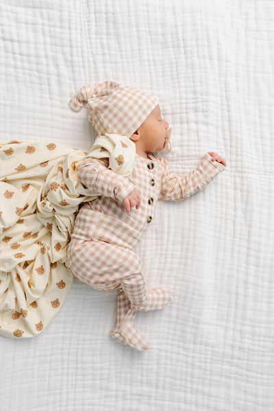 A baby lying on a white muslin blanket, wearing a beige and white gingham footed romper, matching beanie hat, and a cream muslin wrap with small brown teddy bear prints.