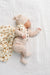 A baby lying on a white muslin blanket, wearing a beige and white gingham footed romper, matching beanie hat, and a cream muslin wrap with small brown teddy bear prints.