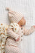 A sleeping newborn baby wearing a peach and cream gingham long-sleeve romper, matching pointed hat, and a cream blanket with teddy bear prints, lying on a white quilted blanket.