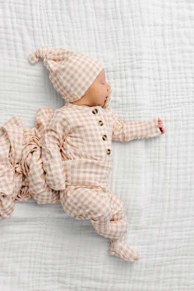 A sleeping newborn baby wearing a matching beige and white gingham footed sleeper and hat, lying on a white textured muslin blanket.