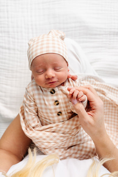 A sleeping newborn baby wrapped in a soft beige and white gingham swaddle blanket and matching hat, held by an adult's hand against a white textured background.