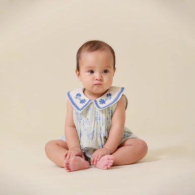 Baby sitting on a beige background wearing a floral dress with a blue collar.