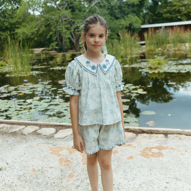 Young girl in a light blue floral outfit standing by a pond with greenery in the background