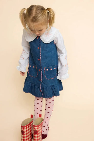A young girl with blonde pigtails wearing a blue denim pinafore dress over a white long-sleeve shirt, pink heart-print tights, and red-and-white gingham rain boots, standing against a solid pale peach background.