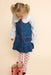 A young girl with blonde pigtails wearing a blue denim pinafore dress over a white long-sleeve shirt, pink heart-print tights, and red-and-white gingham rain boots, standing against a solid pale peach background.
