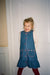 A young girl with blonde hair stands in front of a white paneled door wearing a blue denim pinafore dress over a cream patterned long-sleeve shirt, with dark red tights.