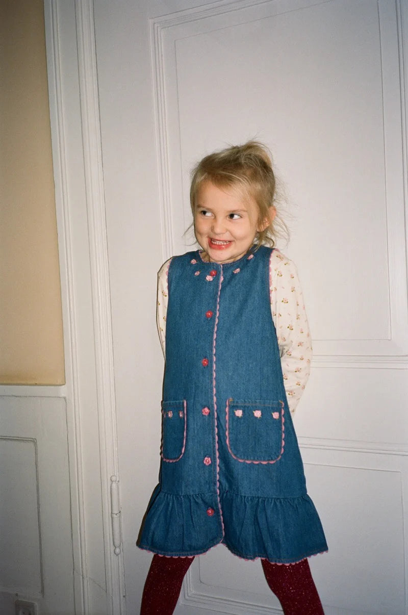 A young girl with blonde hair stands in front of a white paneled door wearing a blue denim pinafore dress over a cream patterned long-sleeve shirt, with dark red tights.
