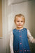 A young girl with blonde hair stands in front of a white paneled door, wearing a blue denim pinafore dress over a white long-sleeve shirt with small pink floral prints.