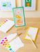Children's art supplies including a drawing of planets and a rocket on an easel, with color swatches and brushes on a wooden surface.
