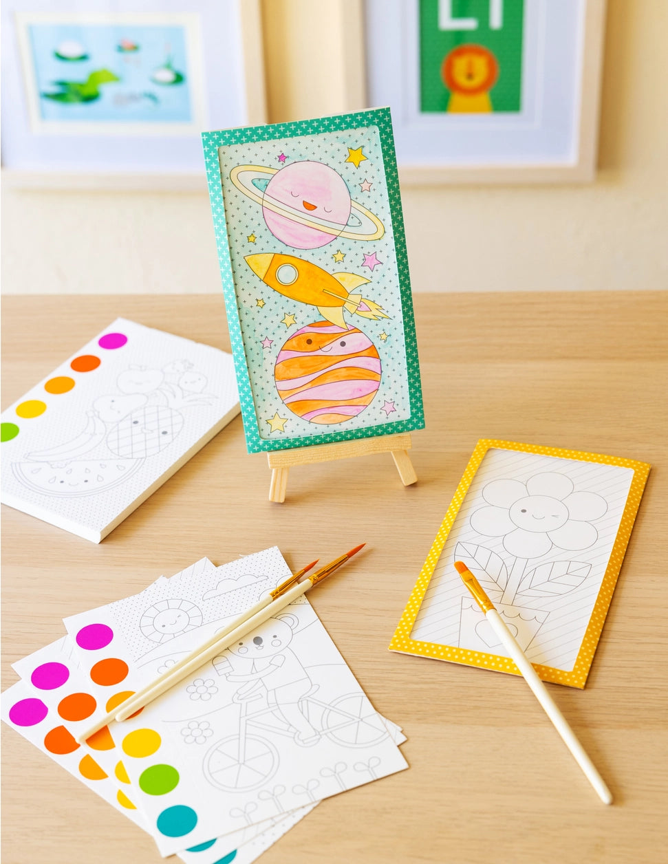 Children's art supplies including a drawing of planets and a rocket on an easel, with color swatches and brushes on a wooden surface.