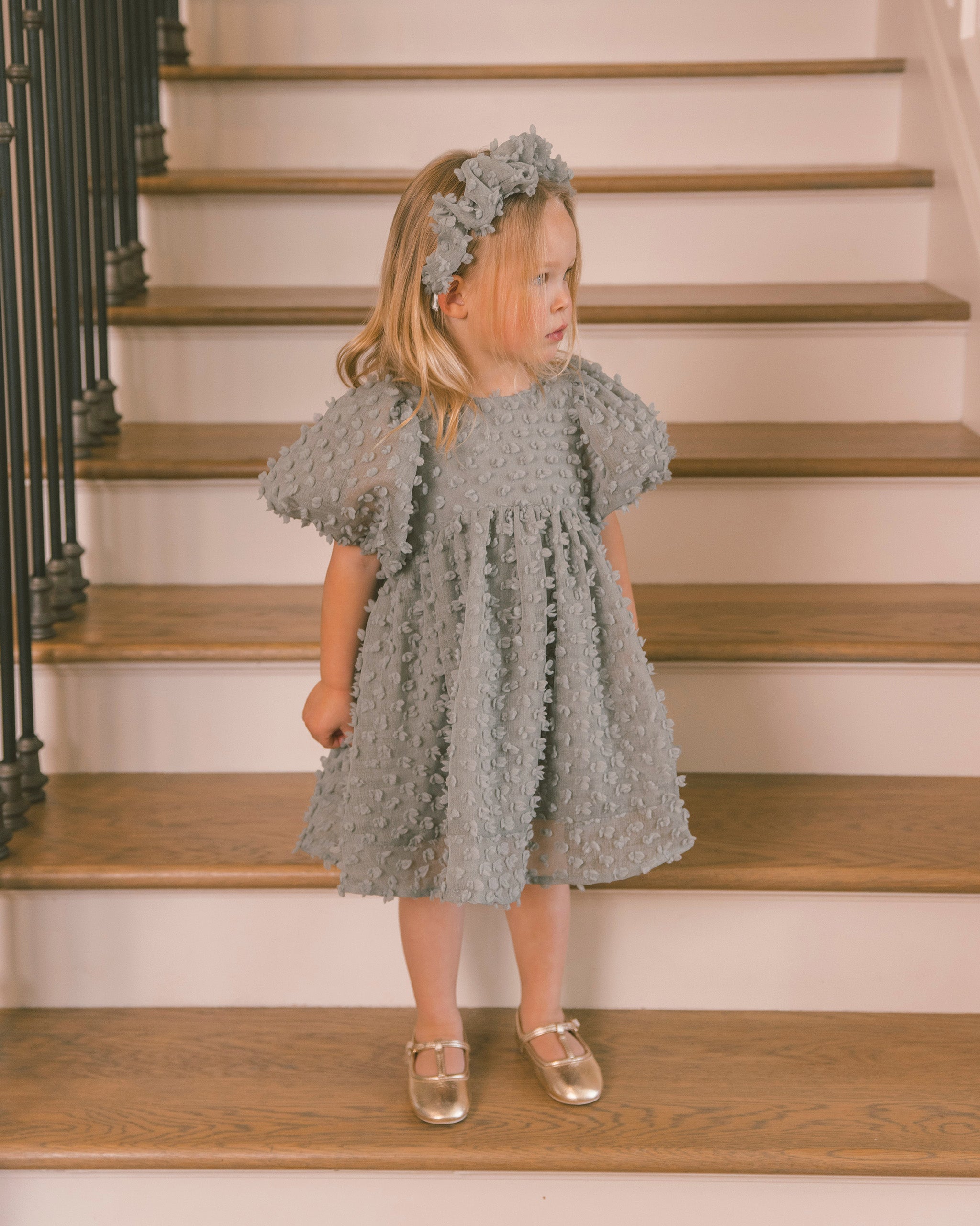 A young girl stands on wooden stairs wearing a textured light gray dress with puff sleeves, a matching floral headband, and metallic gold shoes.