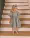 A young girl stands on wooden stairs wearing a textured light gray dress with puff sleeves, a matching floral headband, and metallic gold shoes.