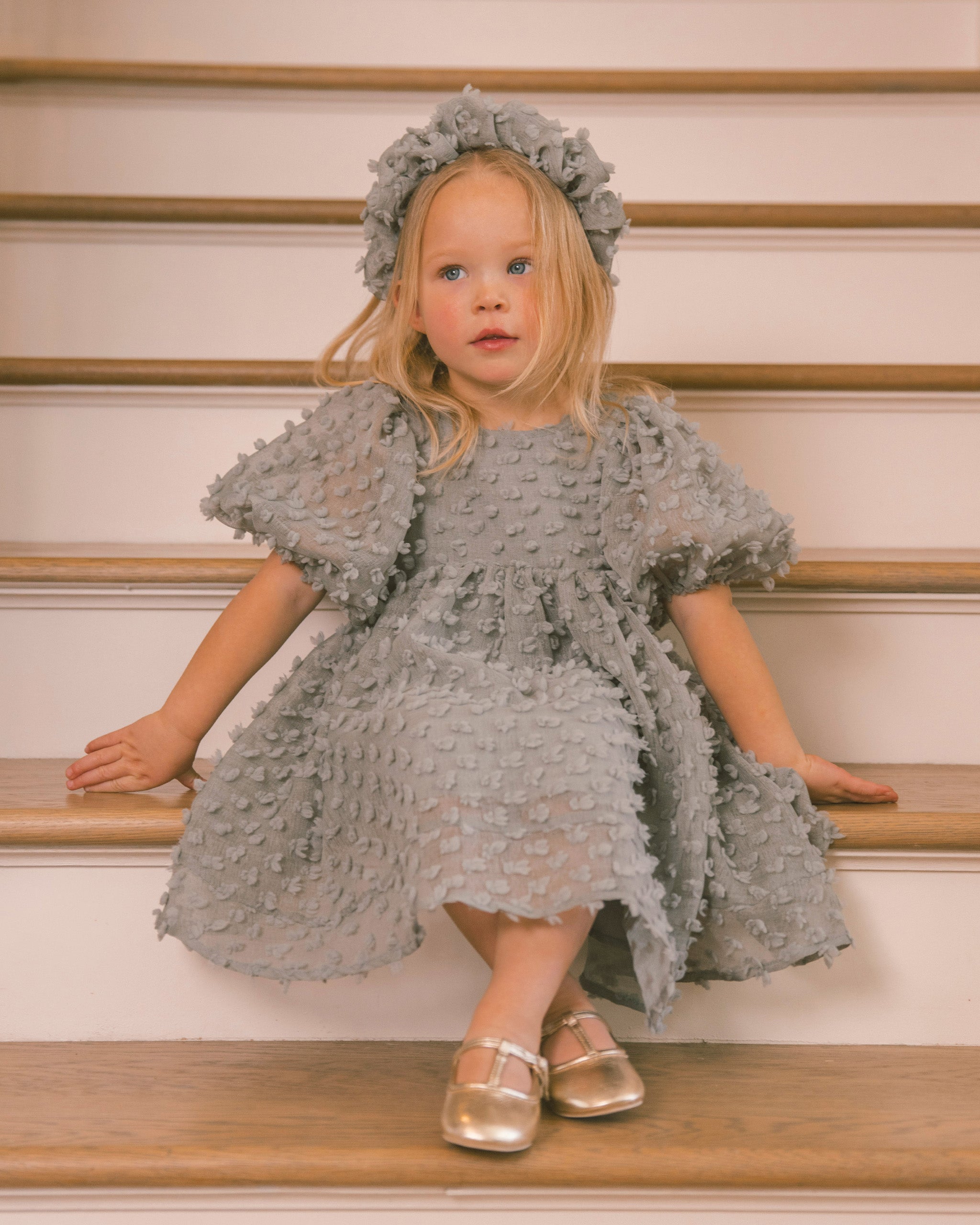A young girl with blonde hair sits on wooden stairs wearing a textured light gray dress with puff sleeves, a matching headband, and metallic gold shoes.