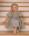 A young girl with blonde hair sits on wooden stairs wearing a textured light gray dress with puff sleeves, a matching headband, and metallic gold shoes.