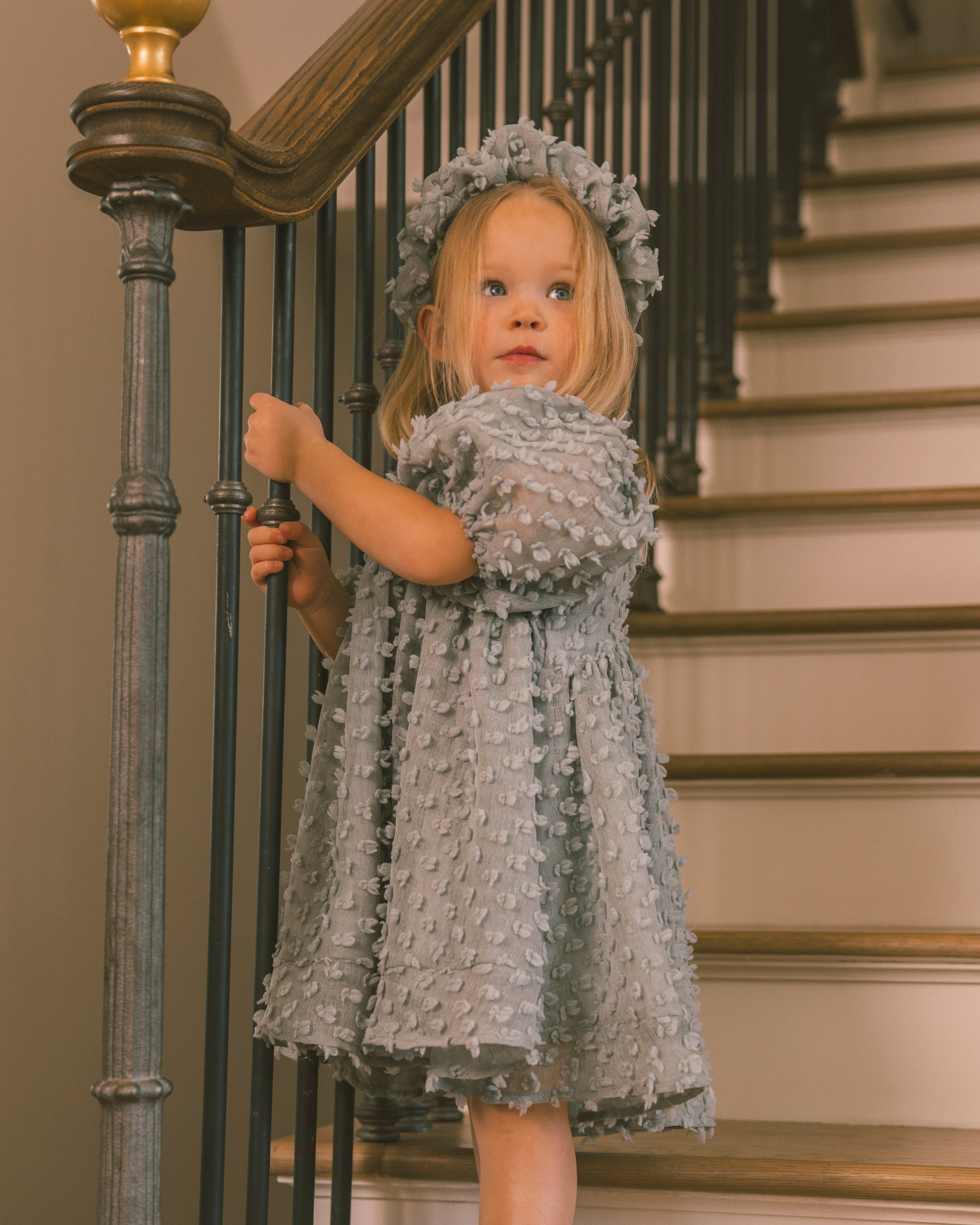 A young girl wearing a textured light gray dress and matching flower crown stands on an indoor staircase, holding the banister.