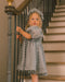 A young girl wearing a textured light gray dress and matching flower crown stands on an indoor staircase, holding the banister.