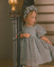 A young girl wearing a textured light gray dress and matching flower crown stands on a wooden staircase next to a banister.