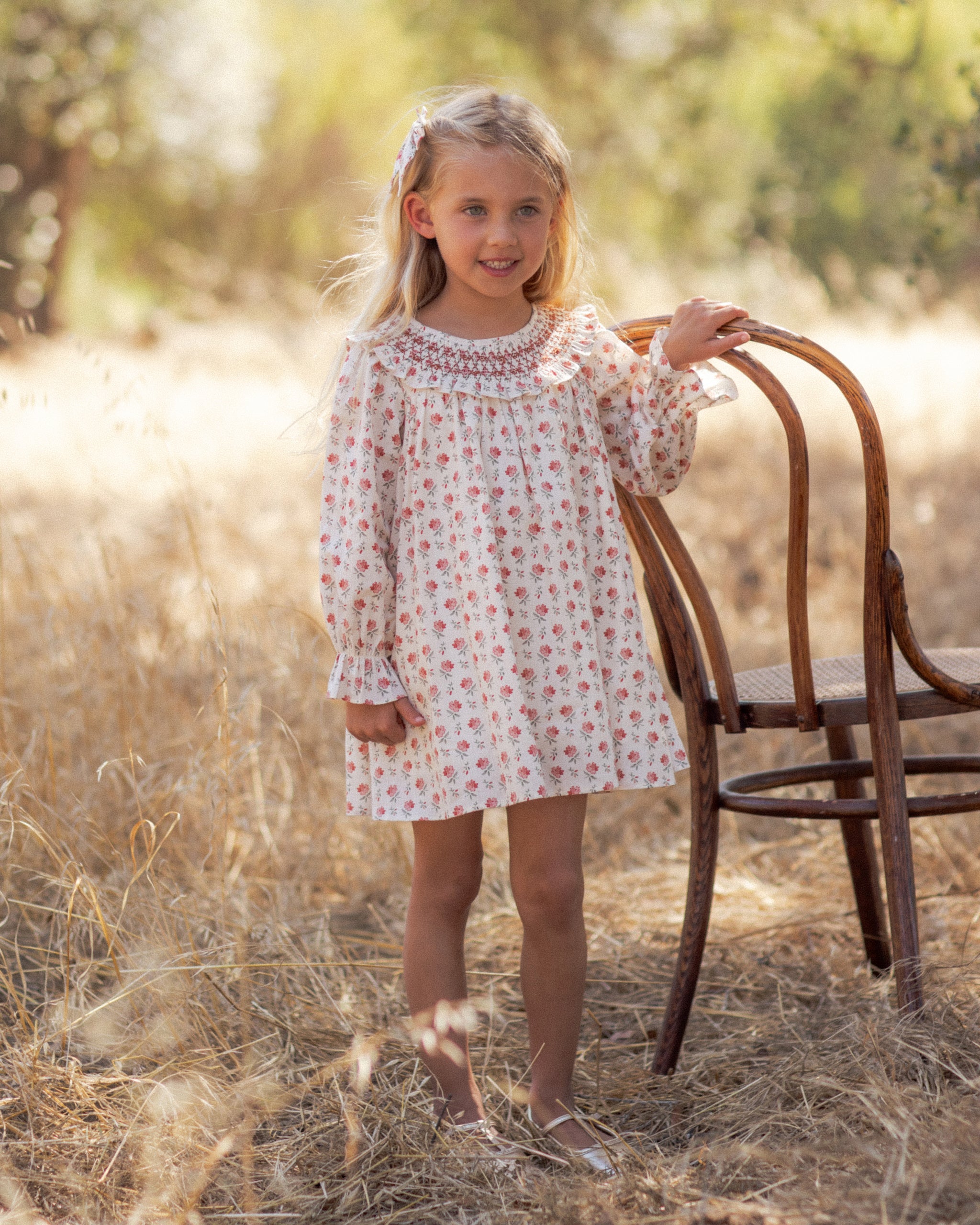 A girl in an ivory floral dress with a smocked collar and long puff sleeves, standing in a golden meadow beside a wooden chair.