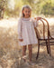 A girl in an ivory floral dress with a smocked collar and long puff sleeves, standing in a golden meadow beside a wooden chair.