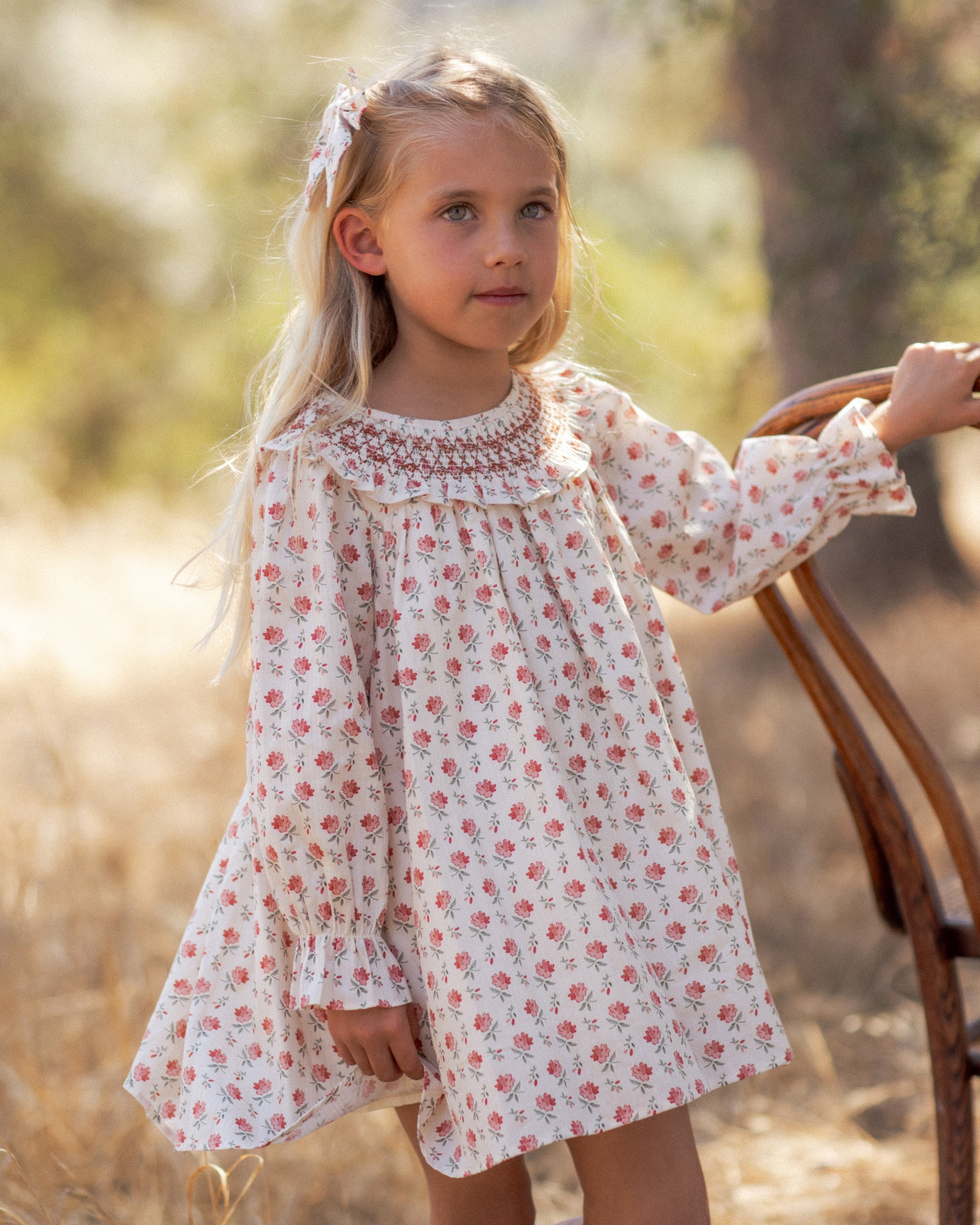 Close-up of a girl in an ivory floral dress with a smocked ruffled collar and puff sleeves, leaning against a wooden chair in a sun-drenched field