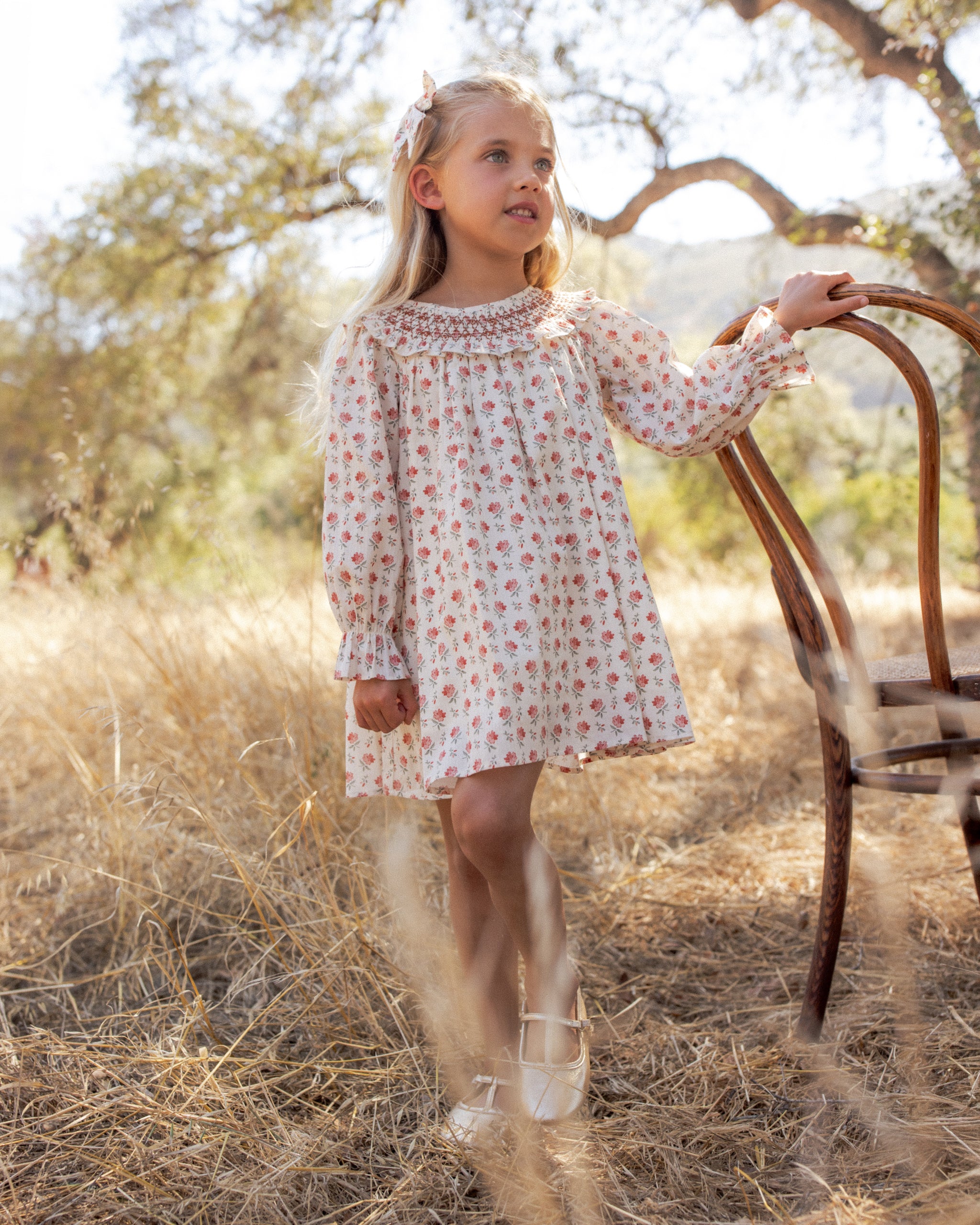 A girl in an ivory floral dress with a smocked collar and puff sleeves, standing outdoors beside a rustic wooden chair with trees in the background.