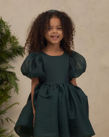 Young girl wearing a dark green puff sleeve dress with a gathered waist and full skirt while smiling indoors beside greenery