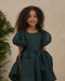 Young girl wearing a dark green puff sleeve dress with a gathered waist and full skirt while smiling indoors beside greenery
