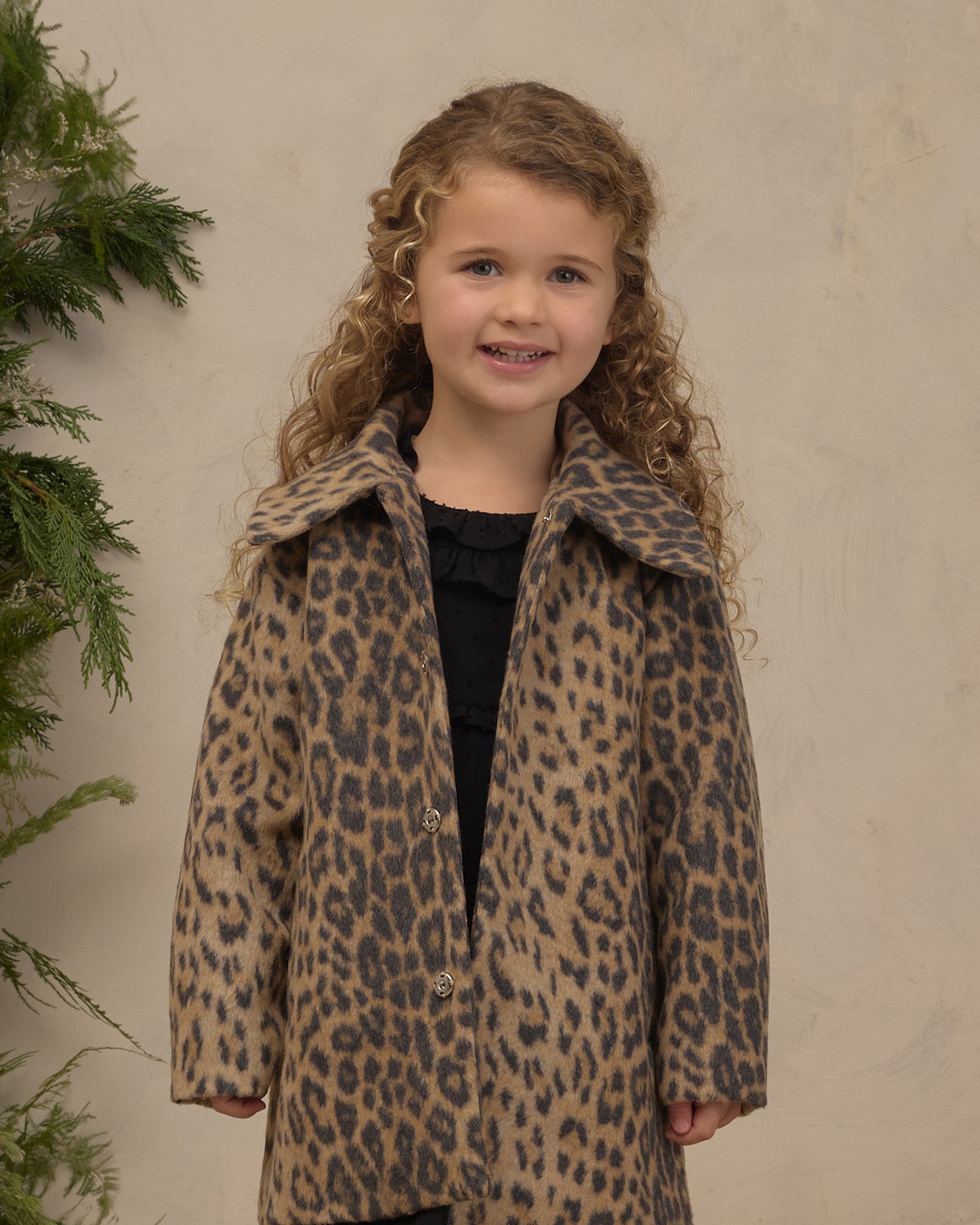 Young girl wearing a Noralee leopard print coat with a full collar and hidden snaps over a black outfit posed indoors beside greenery